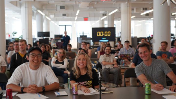 Three people at a table with paper and pen in hand and countdown clock at zero in the back of full room